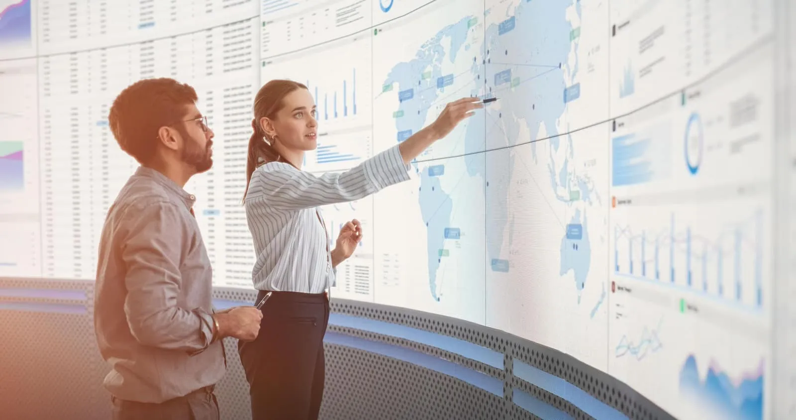Two colleagues examining a curved wall display of sustainability dashboards and a world map, one pointing at a highlighted region.
