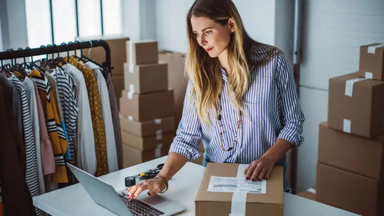 A small business owner packing an order on a laptop next to shipping boxes